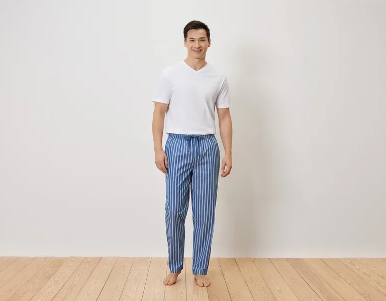 Un homme se tient pieds nus en pantalon de pyjama rayé bleu et blanc et en t-shirt blanc devant un mur blanc.