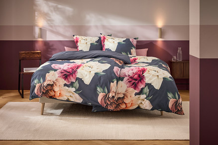 Un lit avec une parure de lit gris foncé à grands motifs floraux roses, blancs et oranges se trouve dans une chambre aux murs bicolores et aux meubles en bois.