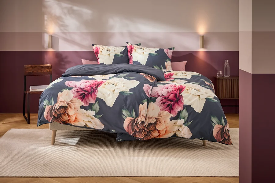 Un lit avec une parure de lit gris foncé à grands motifs floraux roses, blancs et oranges se trouve dans une chambre aux murs bicolores et aux meubles en bois.