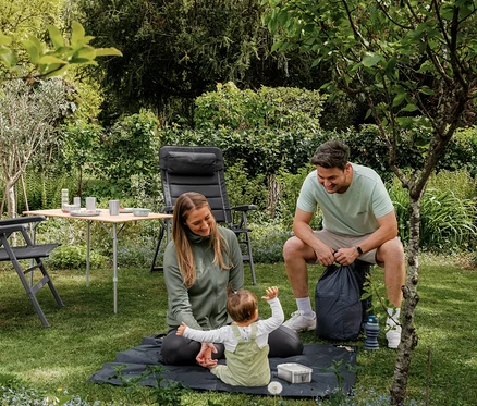 Femme en veste fonctionnelle verte et bébé assis sur une mini-couverture de pique-nique gris foncé dans le jardin, un homme souriant à côté.