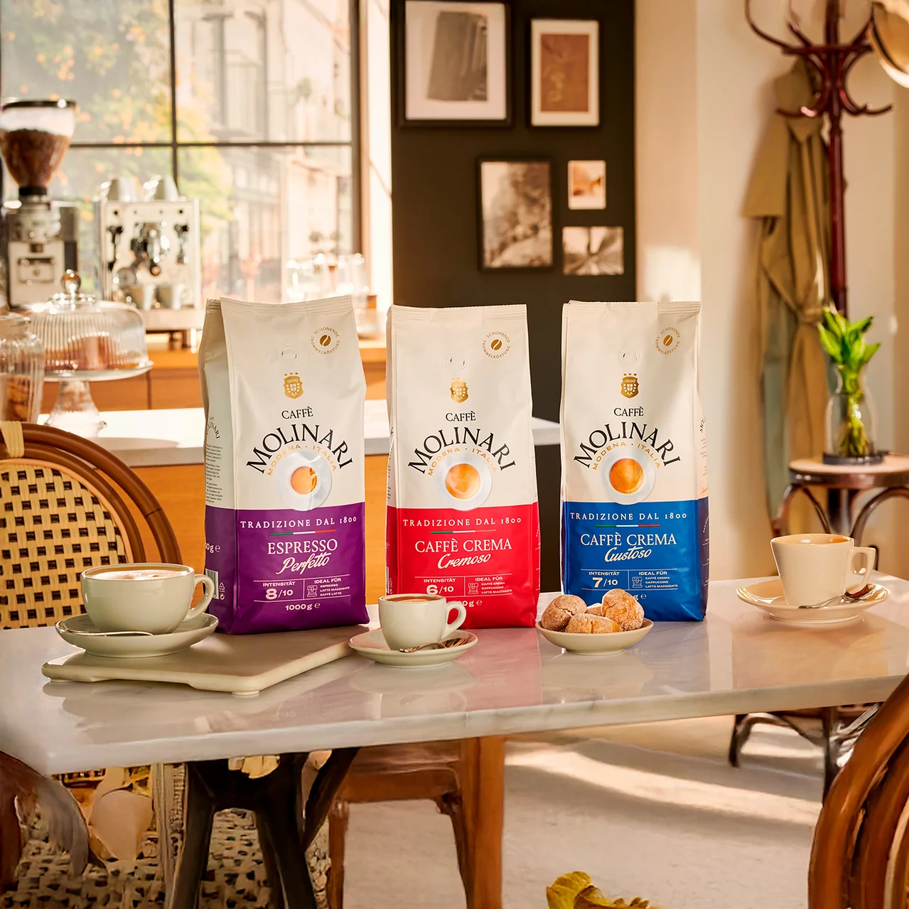 Trois paquets de café Molinari sont posés sur une table à côté de tasses et d'assiettes de pâtisseries.