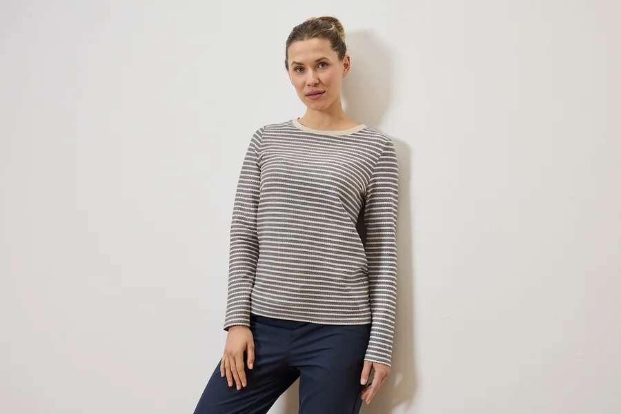 Une femme portant un chignon porte un haut à manches longues rayé gris et blanc et un pantalon bleu marine devant un mur blanc.