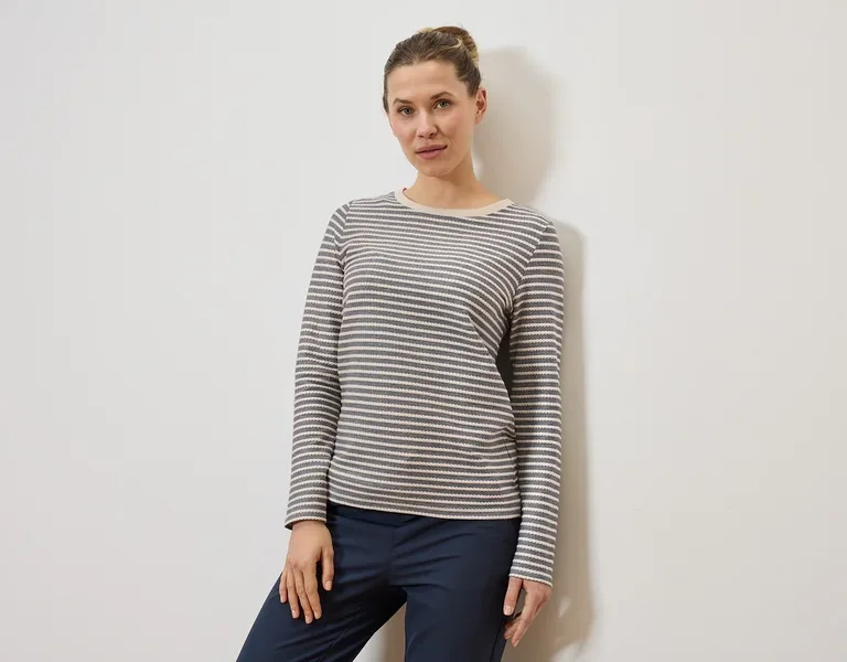 Une femme portant un chignon porte un haut à manches longues rayé gris et blanc et un pantalon bleu marine devant un mur blanc.