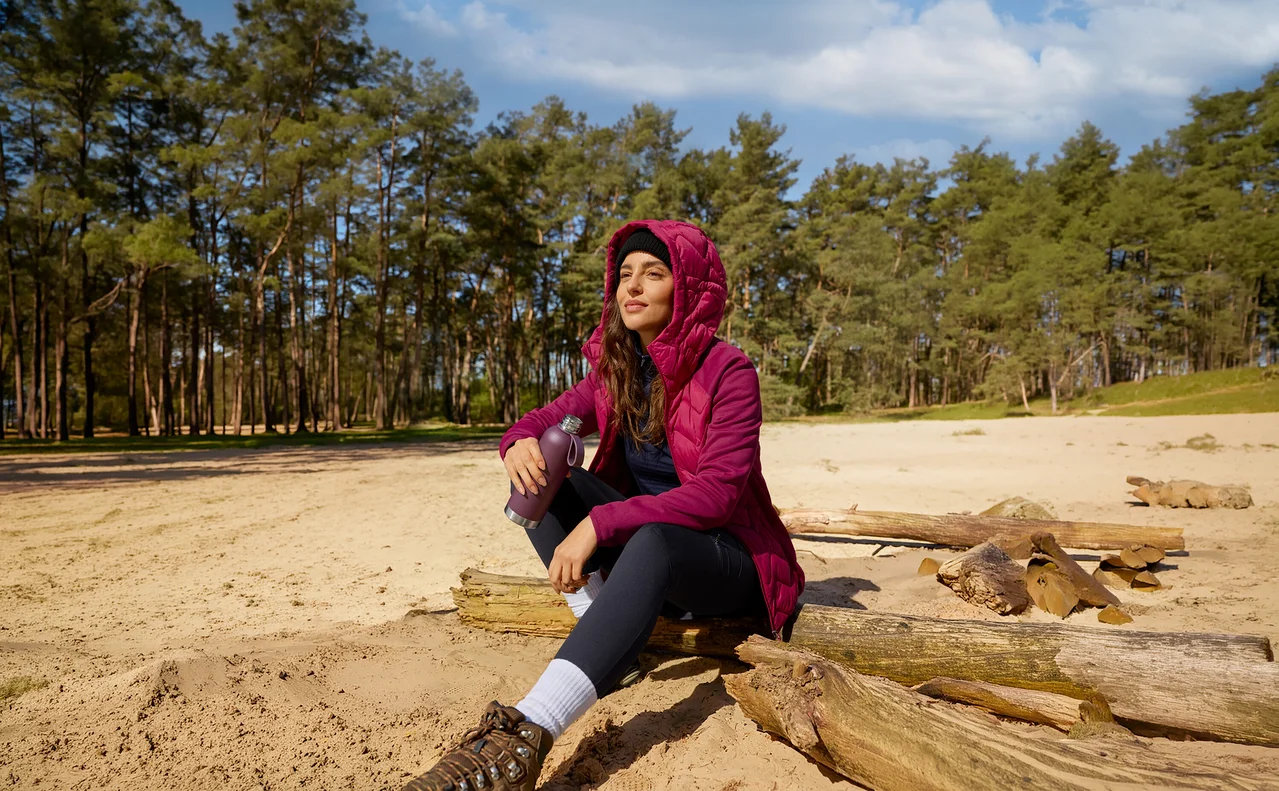 Une femme en veste magenta est assise sur une bûche dans le sable, tenant une gourde de sport et regardant le ciel.
