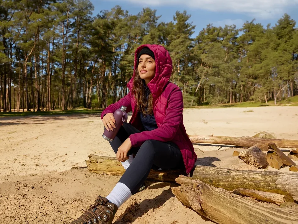 Une femme en veste magenta est assise sur une bûche dans le sable, tenant une gourde de sport et regardant le ciel.
