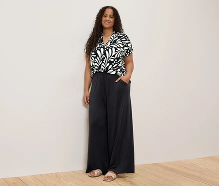 Une femme aux cheveux bouclés, portant un haut à motifs noirs et blancs et un pantalon ample foncé, se tient devant un mur.