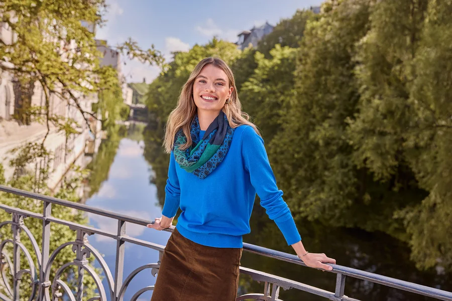 Femme souriante en haut bleu, jupe marron et écharpe à motifs, portant des Créoles doubles en argent 925 rhodié, appuyée sur un pont au-dessus d'une rivière.