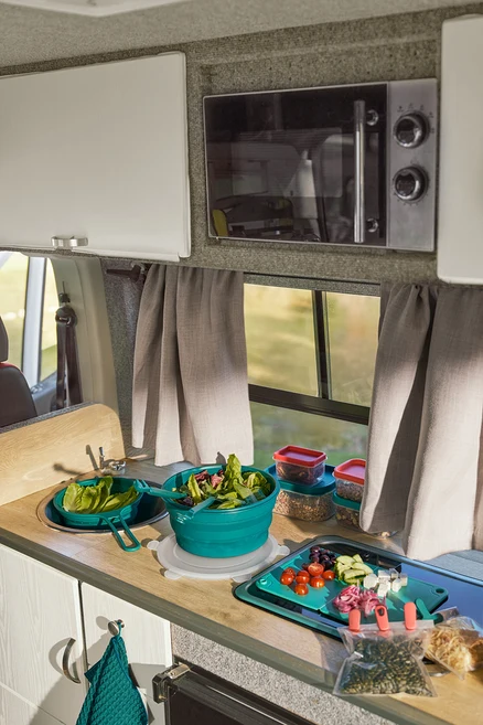 Comptoir de cuisine de camping-car avec une passoire peu encombrante dans l'évier, un saladier pliable avec de la salade et une spatule-pince 2-en-1. Des ingrédients coupés sont sur la planche à découper pliable. À côté, deux clips d'arôme et deux boîtes de conservation. Un micro-ondes est au-dessus de la fenêtre.