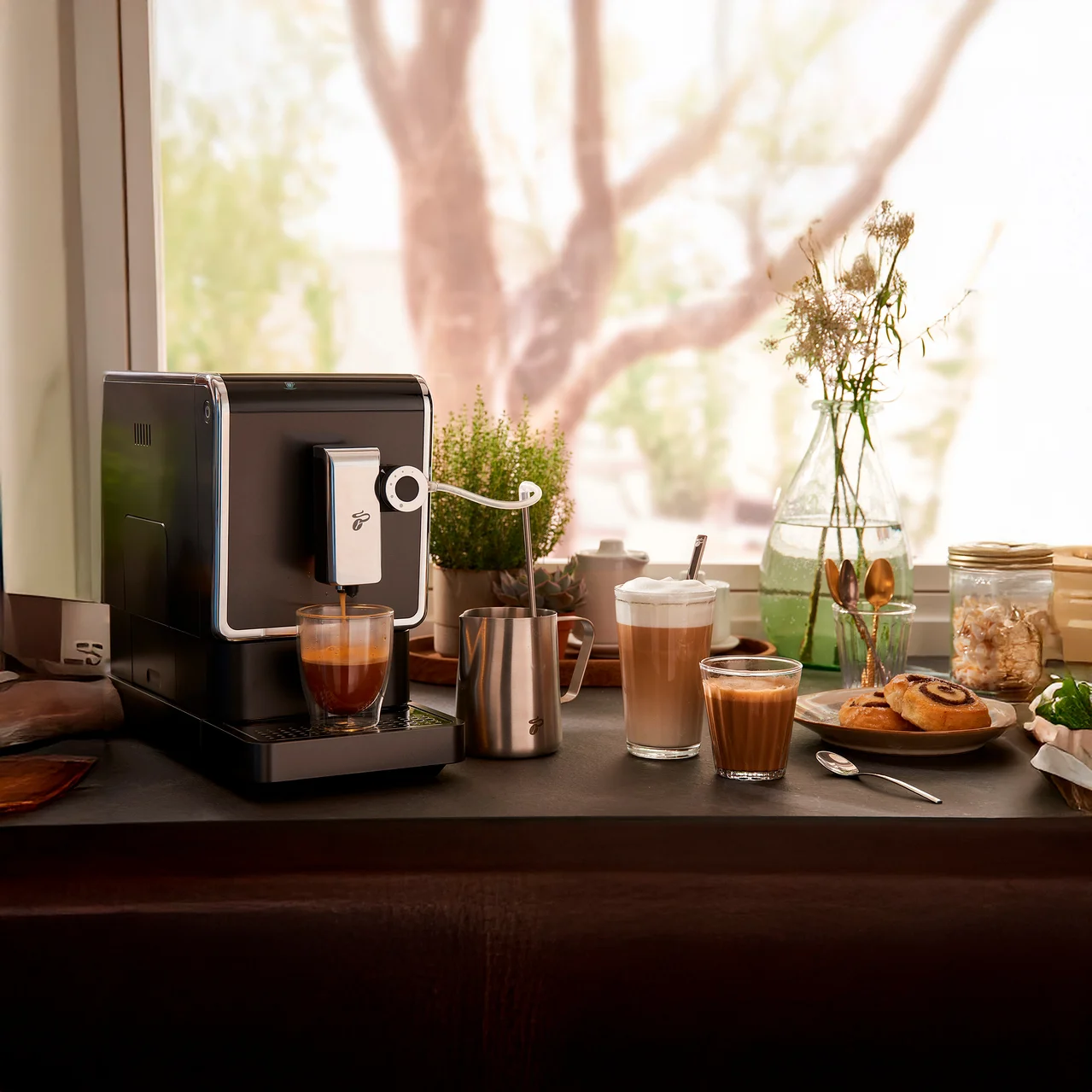 Machine à café préparant du café dans un verre. Sur la table, un mousseur à lait, un latte macchiato et des pâtisseries.