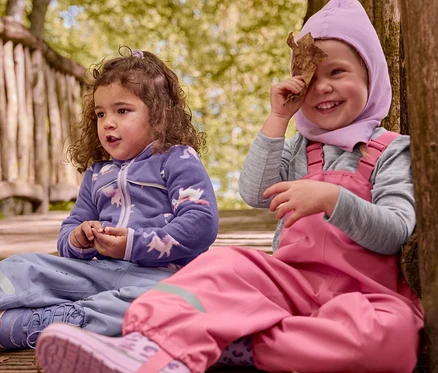 Deux enfants sont assis sur un chemin en bois. L'un porte des bottines thermiques violettes pour enfant et un haut licorne violet. L'autre, en salopette rose et cagoule rose, avec des bottes de pluie roses pour enfant à motif léopard, tient une feuille devant son œil.