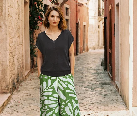 Une femme aux cheveux courts et bruns se tient dans une ruelle, vêtue d'un haut noir à col en V, d'un pantalon vert à motif de feuilles blanches et de boucles d'oreilles créoles.