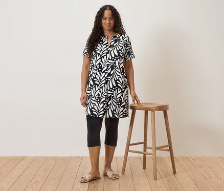 Une femme aux cheveux bouclés portant une robe noire et blanche, des leggings et des sandales se tient à côté d'un tabouret.
