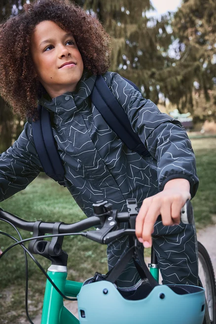 Enfant aux cheveux bouclés, veste foncée à motifs et sac à dos, debout à côté d'un vélo vert, un casque bleu clair accroché au guidon, regarde vers le haut.