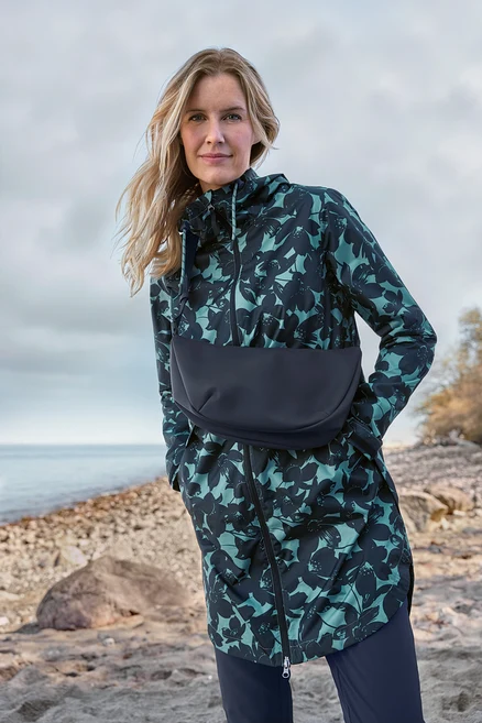 Femme aux cheveux blonds portant une veste à capuche à motif floral vert sarcelle foncé et noir avec un empiècement noir, debout sur une plage rocheuse au bord de la mer.