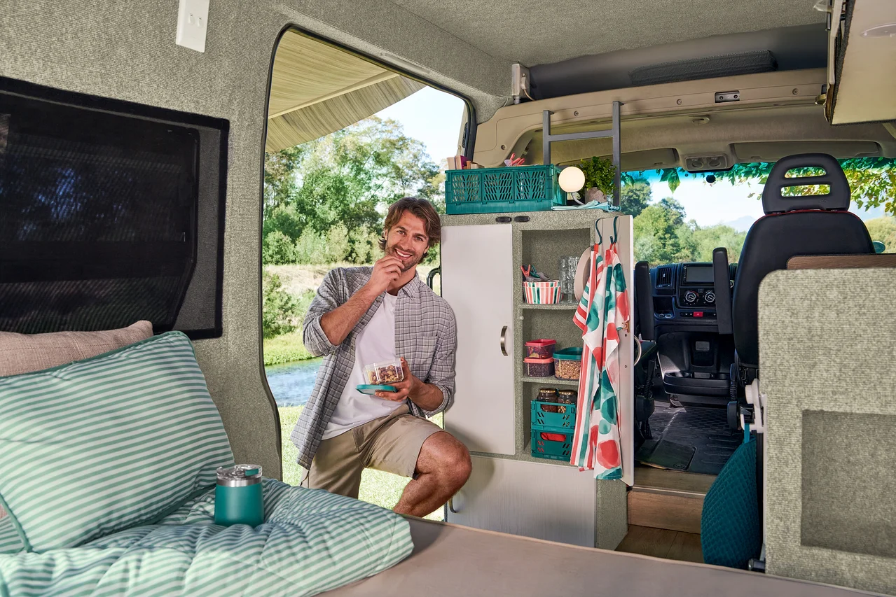 Un homme souriant se tient dans l'embrasure d'un camping-car, tenant une assiette de gâteau et regardant une rivière et des arbres. À gauche, un lit avec un kit de literie de voyage rayé vert et blanc et un thermos turquoise est visible. Derrière lui, une armoire avec des étagères contient divers objets.
