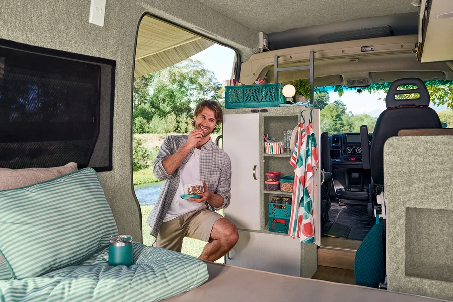 Un homme souriant se tient dans l'embrasure d'un camping-car, tenant une assiette de gâteau et regardant une rivière et des arbres. À gauche, un lit avec un kit de literie de voyage rayé vert et blanc et un thermos turquoise est visible. Derrière lui, une armoire avec des étagères contient divers objets.