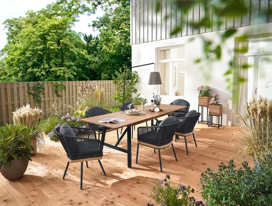 Sur une terrasse en bois se trouvent une table de jardin à rallonges avec structure rustique d'environ 200 à 250 cm, des chaises de jardin pivotantes avec tressage textile et des chaises de jardin avec tressage textile. On voit aussi 2 colonnes à plantes « Tinus » et une lanterne solaire à accu avec support de table.