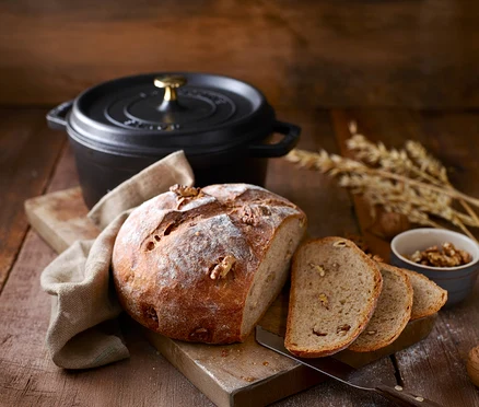 Une miche de pain aux noix et des tranches de pain sur une planche à découper avec une cocotte STAUB »Cocotte 24« noire en arrière-plan.