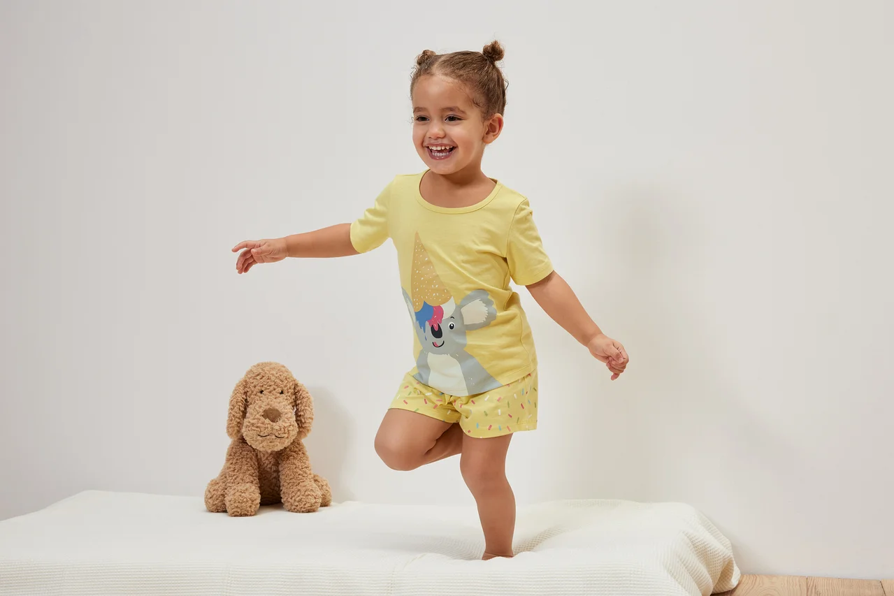Une fillette souriante se tient debout sur un lit à côté d'un chien en peluche et porte un pyjama court jaune pour enfant, koala.
