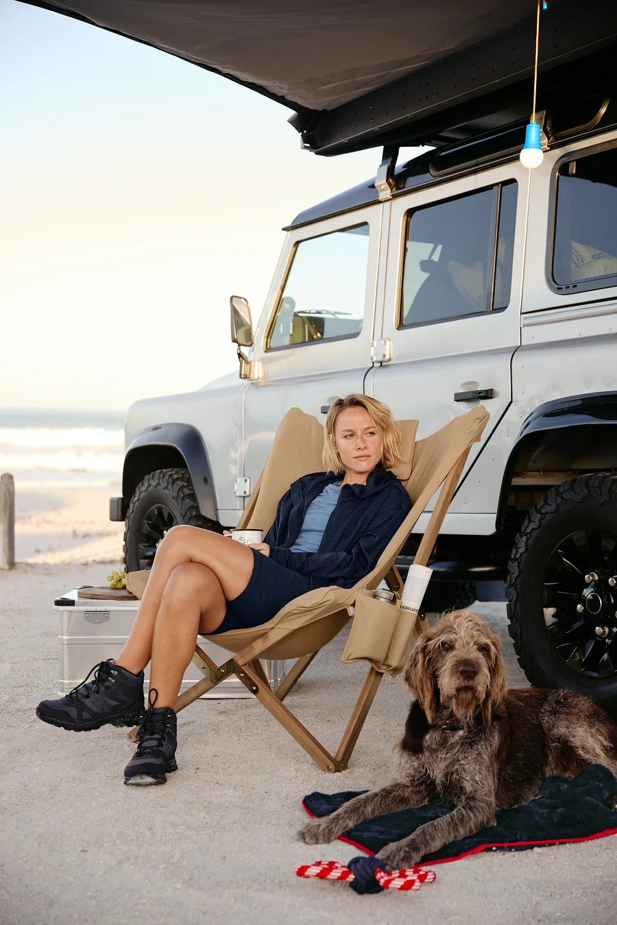 Une femme assise dans un fauteuil relax avec un mug, un chien est allongé à côté sur une couverture moelleuse pour chien avec un jouet. Une lampe de camping à LED, un pantalon fonctionnel 2 en 1 et une veste à capuche sont à l'arrière.