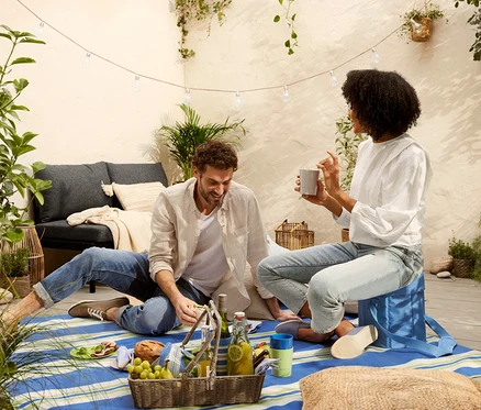 Un homme et une femme sont assis sur une nappe de pique-nique XL. Devant eux se trouvent un sac isotherme 2 en 1 et un panier-plateau contenant de la nourriture.