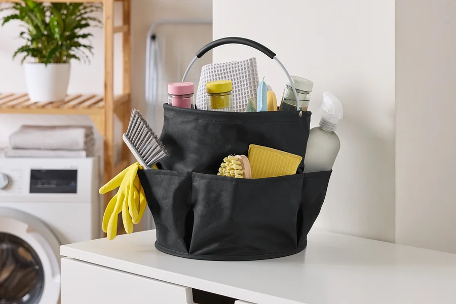 Photo d'un panier de nettoyage noir rempli de produits de nettoyage, incluant des brosses, des sprays et des chiffons, sur une surface blanche.