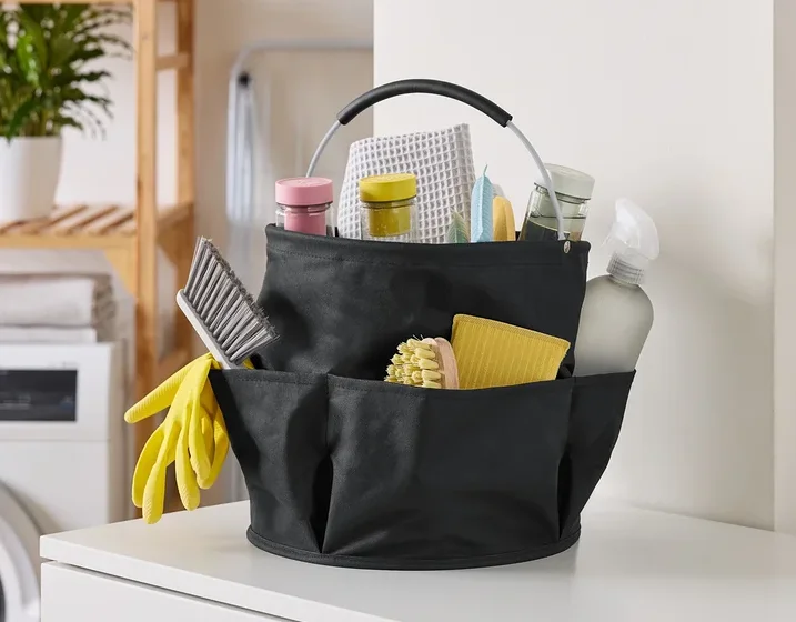Photo d'un panier de nettoyage noir rempli de produits de nettoyage, incluant des brosses, des sprays et des chiffons, sur une surface blanche.