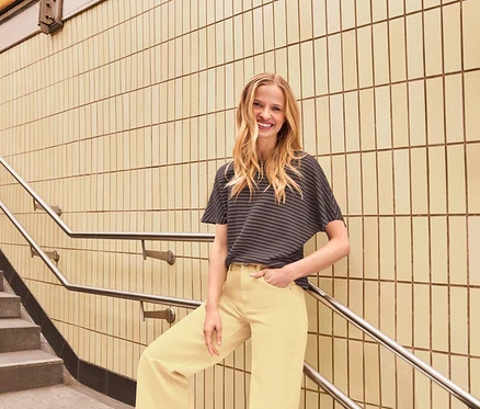 Une femme souriante aux longs cheveux blonds porte un haut à manches courtes à motifs sombres et un pantalon large jaune clair. Elle est appuyée contre une rampe à côté d'un mur carrelé et d'un escalier.