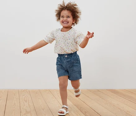 Une petite fille souriante court sur un plancher en bois et porte un short en jean et des sandales.