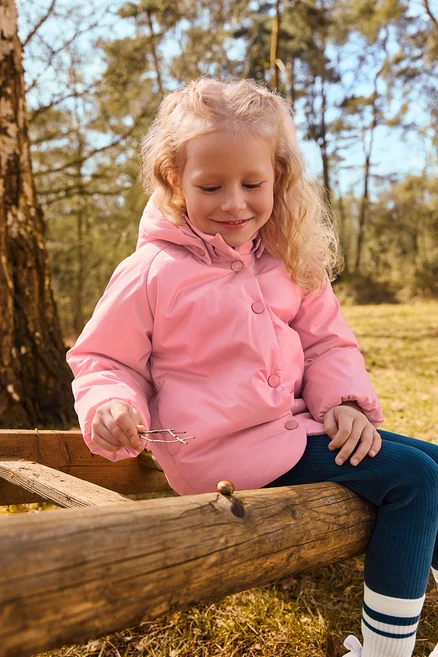 Une petite fille aux cheveux blonds bouclés, vêtue d'une doudoune rose et d'un legging bleu foncé, est assise sur une clôture en bois dans une forêt, tenant de petites brindilles et regardant la clôture.