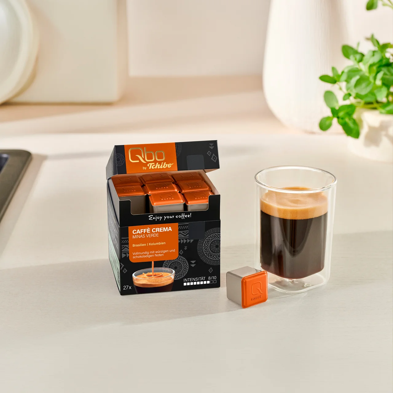 Paquet ouvert de capsules de café Qbo by Tchibo Caffè Crema Minas Verde avec 27 capsules orange, à côté d'un verre de café préparé et d'une capsule individuelle sur un comptoir.
