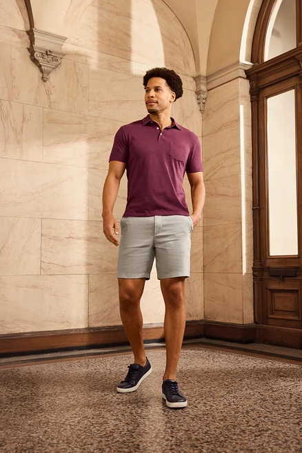Un homme aux cheveux bouclés porte un polo bordeaux, un short et des chaussures bleu marine. Il se tient devant un mur orné et une porte en bois.