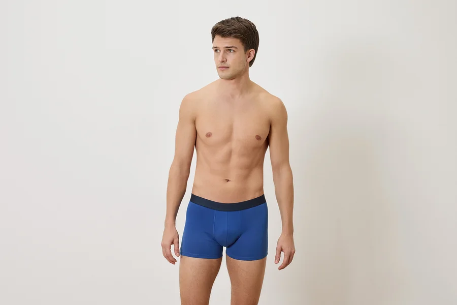 Homme torse nu portant un boxer bleu sur un fond blanc.