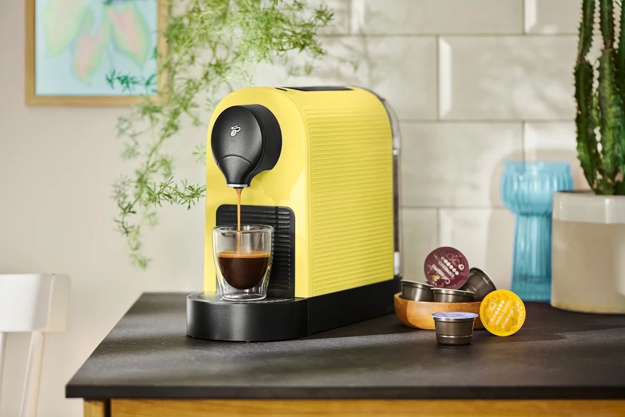 Une machine à café jaune remplit une tasse de café, des capsules sont à côté.