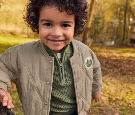 Un jeune garçon souriant aux cheveux bouclés se tient près d'un arbre dans une forêt ensoleillée, portant une veste matelassée beige sur un pull côtelé vert.
