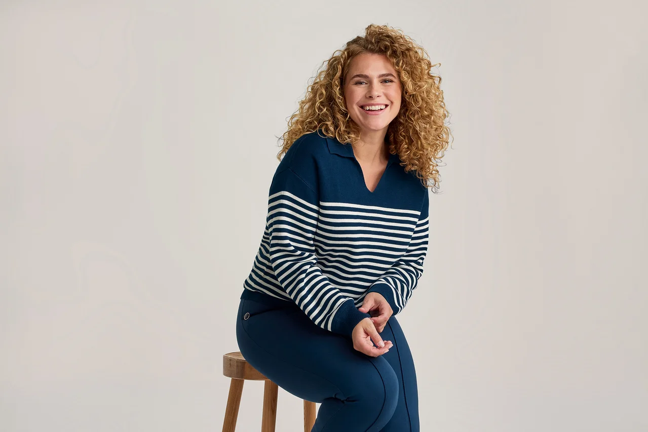Femme souriante aux cheveux bouclés portant un pull rayé bleu marine et blanc et un pantalon bleu marine, assise sur un tabouret en bois.