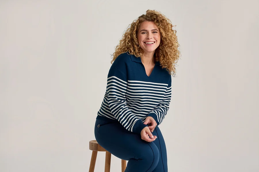 Femme souriante aux cheveux bouclés portant un pull rayé bleu marine et blanc et un pantalon bleu marine, assise sur un tabouret en bois.