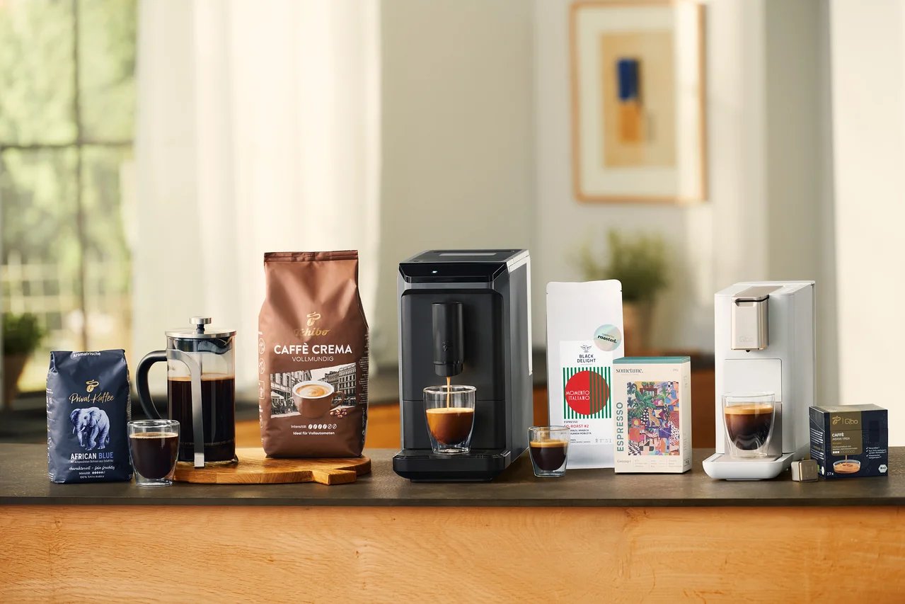 Divers produits de café sur un comptoir : un sachet bleu de Privat Kaffee African Blue en grains, une cafetière à piston en verre avec du café, un verre à double paroi pour café crème, un sachet marron de Caffè Crema Vollmundig en grains, une machine à café entièrement automatique Esperto mini grise versant du café dans un verre à double paroi pour café crème, un petit verre à double paroi pour café crème, un sachet blanc de café, une boîte colorée de café, une machine à café Qbo ICONIC Light Gold blanche versant du café dans un verre à double paroi pour café crème, et une boîte noire de capsules Qbo Caffè Jabana Yirga.