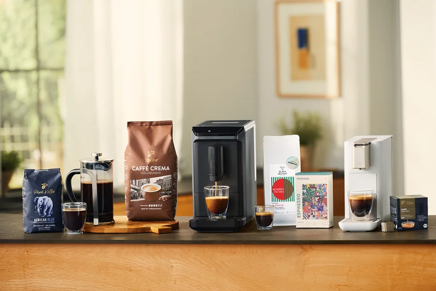 Divers produits de café sur un comptoir : un sachet bleu de Privat Kaffee African Blue en grains, une cafetière à piston en verre avec du café, un verre à double paroi pour café crème, un sachet marron de Caffè Crema Vollmundig en grains, une machine à café entièrement automatique Esperto mini grise versant du café dans un verre à double paroi pour café crème, un petit verre à double paroi pour café crème, un sachet blanc de café, une boîte colorée de café, une machine à café Qbo ICONIC Light Gold blanche versant du café dans un verre à double paroi pour café crème, et une boîte noire de capsules Qbo Caffè Jabana Yirga.