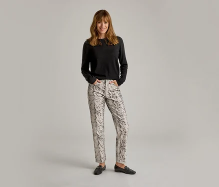 Femme souriante debout, les mains dans les poches, portant un haut noir à manches longues, un pantalon clair à motif serpent et des mocassins noirs.
