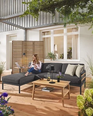 Une femme est assise sur le coin salon de jardin Premium « Elin » avec un paravent flexible en arrière-plan. On aperçoit également un lampadaire d'extérieur à LED sans fil noir et une lampe de table ainsi qu'une table basse de jardin.