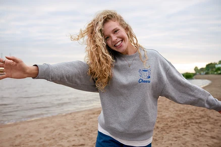 Une femme aux cheveux bouclés portant un sweat-shirt gris avec l'inscription "Cheers" se tient sur la plage, les bras écartés.