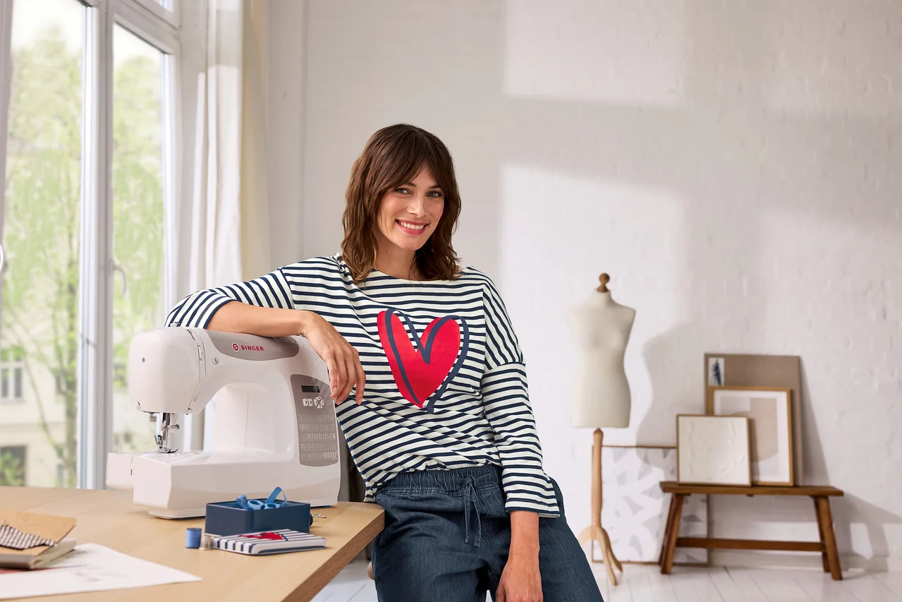 Une femme souriante en t-shirt rayé avec un cœur rouge s'appuie sur une machine à coudre Singer blanche sur une table en bois. À gauche une fenêtre, à droite un mannequin et des tableaux.