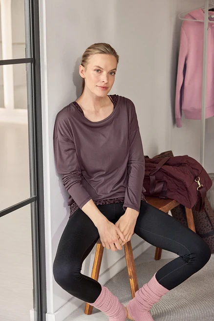 Une femme est assise sur un tabouret, adossée à un mur, vêtue d'un haut à manches longues foncé avec un imprimé léopard, d'un legging noir et de jambières roses.