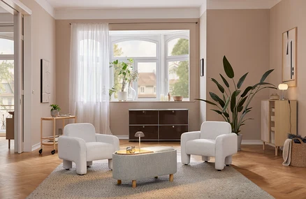 Un salon confortable avec deux fauteuils gris clair et un pouf assorti sur un tapis. Une lampe de table à LED sans fil beige est posée sur une armoire en bois clair à côté d'une grande plante. Un meuble de rangement marron foncé se trouve sous une fenêtre en baie.