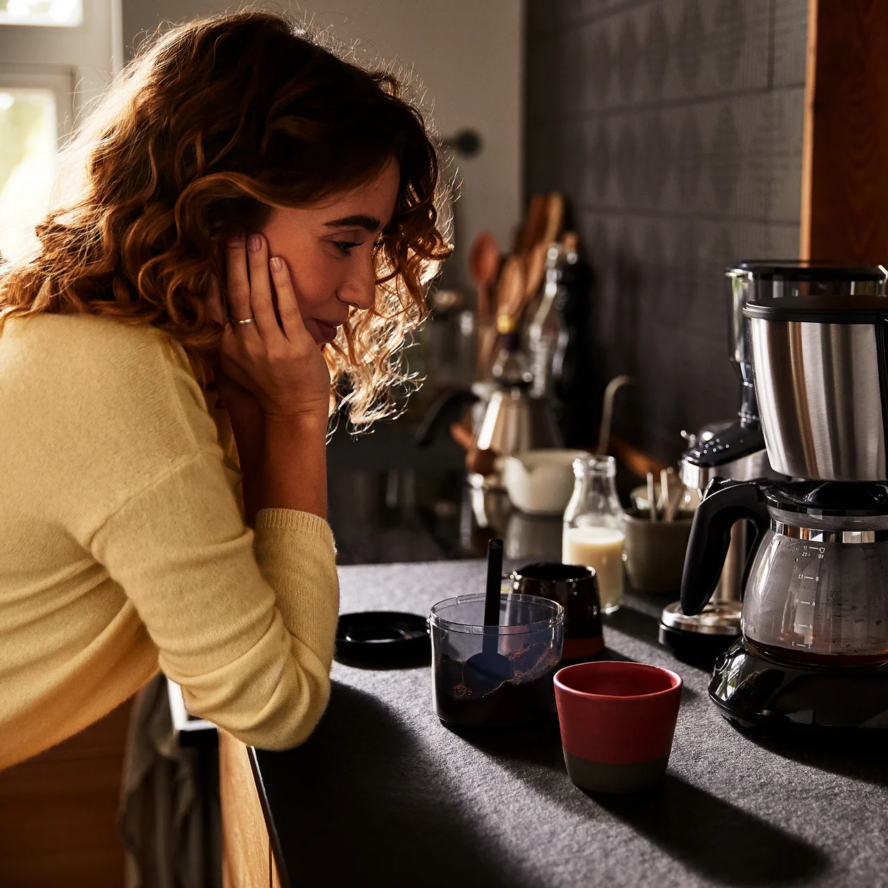 Femme appuyée sur un comptoir de cuisine à côté d'une machine à café, une tasse et un verre.