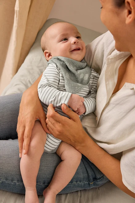 Une mère tient un bébé souriant qui porte un body évolutif pour bébé et un foulard en mousseline pour bébé.