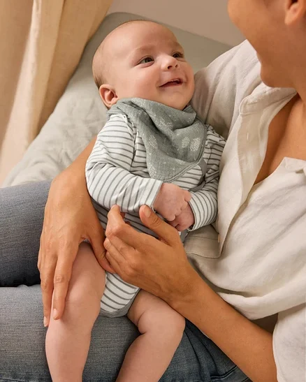 Une mère tient un bébé souriant qui porte un body évolutif pour bébé et un foulard en mousseline pour bébé.