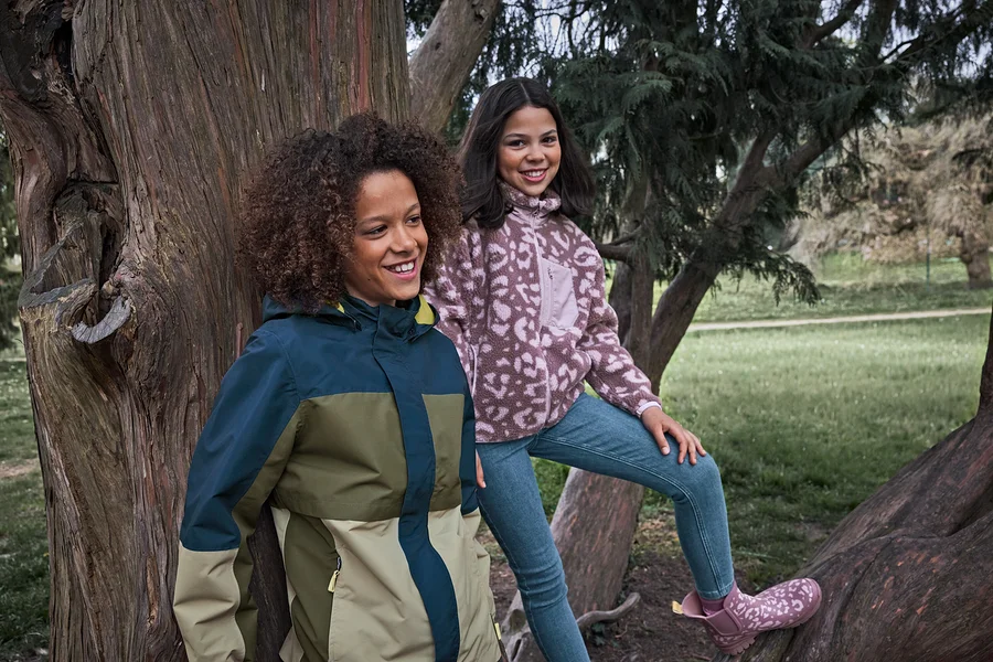 Deux filles souriantes dans un parc, l'une porte une veste multicolore, l'autre est assise sur une branche d'arbre en veste polaire à motifs et Jeans slim pour enfant – Coupe « Lotta ».