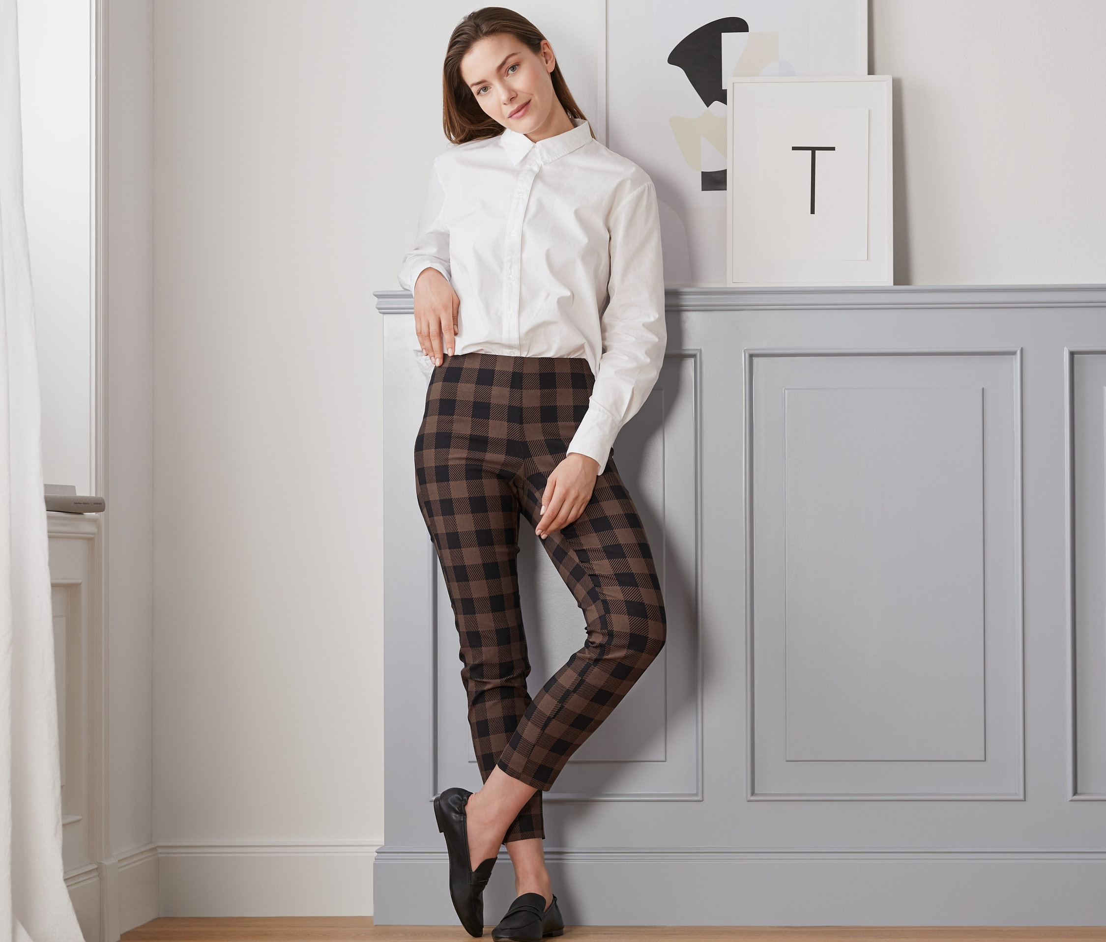 Une femme appuyée contre un mur porte un pantalon stretch à carreaux, une chemise blanche et des mocassins noirs.